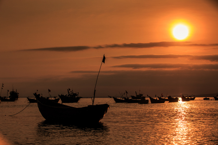 traditional fishing boat with flagstaff in bay and large orange sun above clouds lights silhouettes under dark skyの写真素材