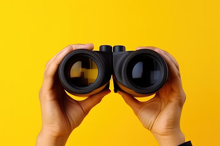 Hand with binoculars on yellow background symbolizes search and explorationの素材