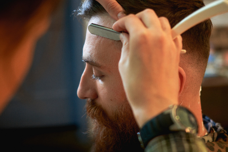 Close up of a hairdressers work for an attractive young blond man at the barber shop. He is doing styling of his beard, shaving it with straight razorの写真素材