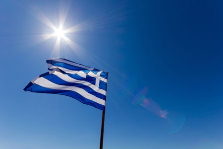 greek flag against the deep blue sky with sun and lens flairの写真素材