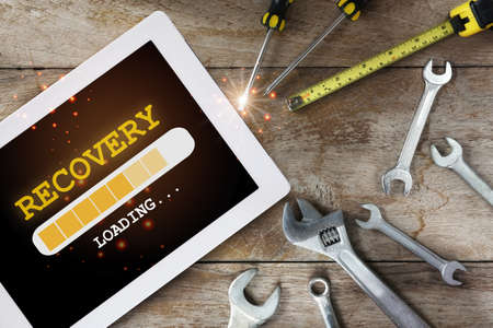 Recovery word loading on digital computer tablet and tools supplies on wooden background. Business crisis management concept and technology transformation ideaの写真素材