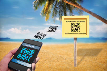 Hand holding smartphone with scanning QR code smart guide screen with guide for visitors sign on tropical beach background. Smart technology concept and qr code quick and easy ideaの写真素材