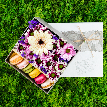 Pink, white, purple flowers and colorful macaroons in a gift box on green background, top view, flatの写真素材