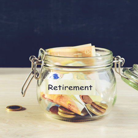 Coins and paper money in a glass jar on wooden table against dark background and word Retirement...