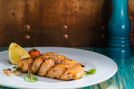 close-up tasty cooked salmon on a white plate with lemon, tomato and bay leaves, on a wooden table. Brown background, blue pepperboxの写真素材