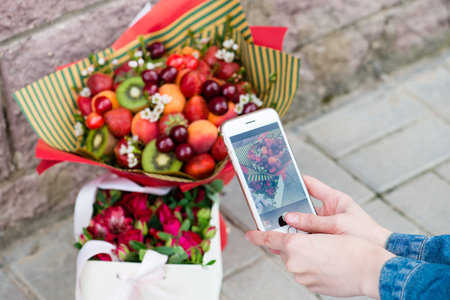 Close-up of a girl hand with a smartphone shoot bouquet On the foreground fruit bouquet and gift box...