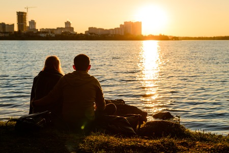 Back view of a man and woman couple sitting by the lake water at sunsetの写真素材