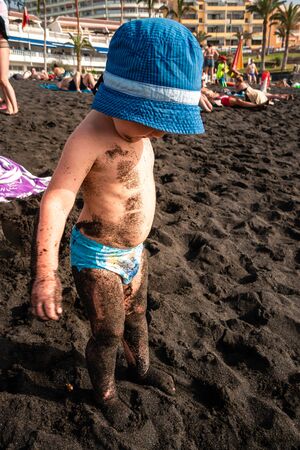 Funny photo of happy baby boy with dirty body. Family travel lifestyle, recreation on summer beach vacation with children. Child boy on the beach smeared in black sand in Tenerifeの写真素材