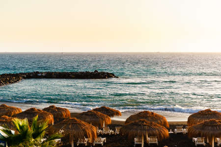 straw beach umbrella with blue sky, sunbeds on the black volcanic beach of La Caletta. Tenerife, Canary Islands, Spain.の写真素材