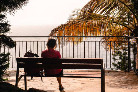 A traveler woman sits on a tropical park under palm trees and enjoys the view to ocean, unknown woman looking at the seaの写真素材