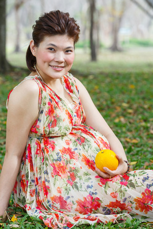 Beautiful asian pregnant woman sit on lawn in public park hold one orange fruit in hand - photo taken 2 days before baby birthの写真素材