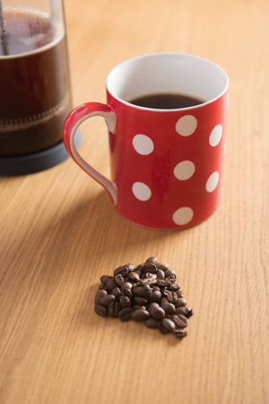 Coffee beans on a wooden table with a freshly brewed cafetiere and red spotty cup in the backgroundの写真素材