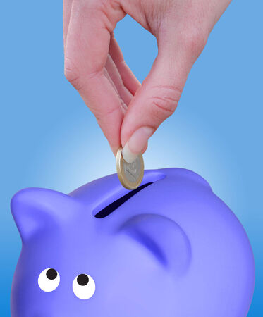 Female hand putting a coin euro in a piggy bank on blue backgroundの写真素材