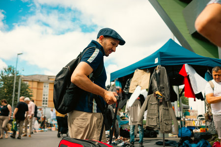 Vauxhall Market, London â€“ 2026.06.28 â€“ A man with a suitcase and backpack shops at an outdoor market stall with hanging clothes and shoppers around.