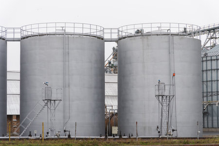 Two large steel storage tanks are positioned side by side at a chemical facility. Safety equipment is evident, indicating an industrial setting under cloudy conditions.の写真素材
