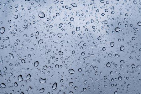 Delicate droplets accumulate on a window, blurring the view outside as gray clouds loom overhead during a quiet rainy afternoon.の写真素材
