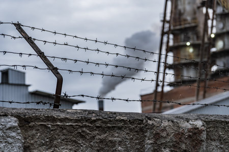 A barbed wire fence stands in the foreground, with a factory in the background exhaling smoke into the gray sky, reflecting an industrial environment.の写真素材