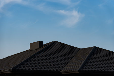 The roof of a suburban house is visible against a bright blue sky with wispy clouds. The textured tiles create a distinct pattern, enhancing the architectural features.の写真素材