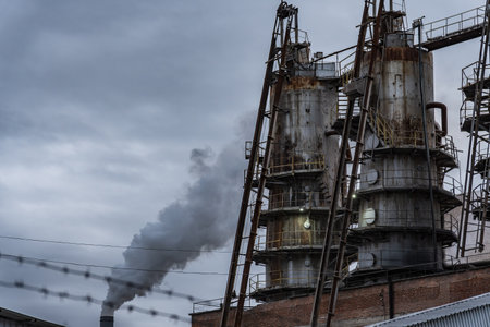 Tall, cylindrical structures with numerous metal platforms and ladders are situated against a gray sky with a thick plume of smoke rising from a nearby chimney.の写真素材