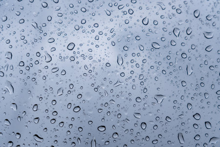 Raindrops cling to a glass surface, distorting the visibility of a gray sky. The droplets vary in...