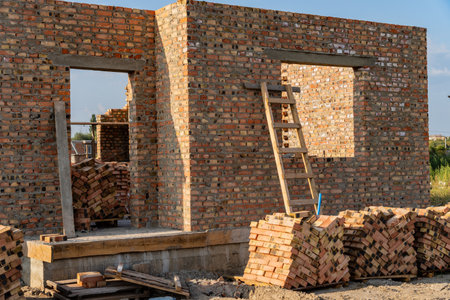 Construction workers are building a brick wall, which stands partially completed. A wooden ladder leans against the structure, and neatly stacked bricks nearby await use.の写真素材