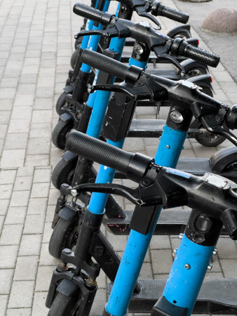 A row of blue scooters are parked on a brick sidewalk. The scooters are all facing the same...