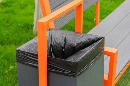 This container, featuring a black bag, is designed for waste disposal and recycling. It is conveniently situated beside a colorful park bench in a lush green space.の写真素材