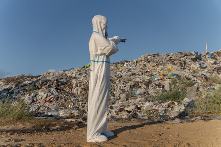A person in protective gear stands at a trash dump, pointing towards a large pile of garbage. The...