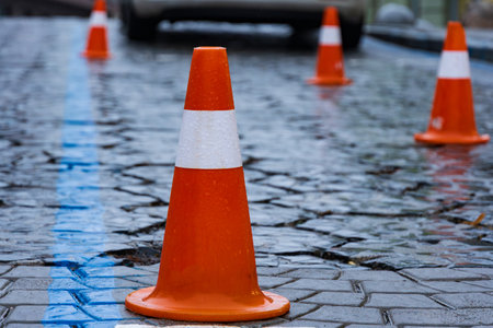 Colorful traffic cones provide clear direction on a wet and glossy street. Rain droplets enhance the environment, reflecting colors and creating patterns on cobblestones.の写真素材