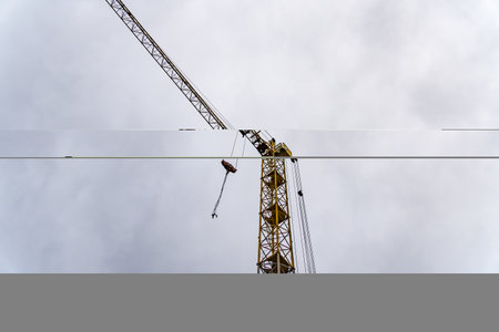 A tall construction crane looms over the skyline, its yellow structure contrasting with the grey clouds. It is actively involved in lifting materials for a new building project.の写真素材