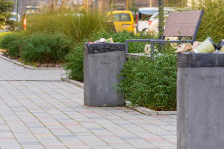 Unkempt garbage containers overflow with trash bags and recycling materials beside a pathway in an urban public area.の写真素材