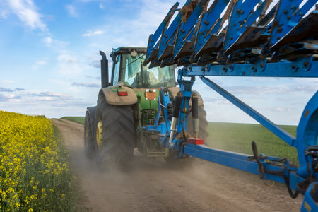 A tractor works through a dirt path, plowing soil beside vibrant yellow canola flowers under a clear...