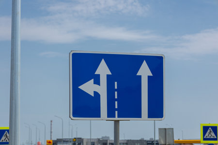 A road sign displays arrows pointing straight and left, guiding drivers at an intersection under a blue sky.の写真素材