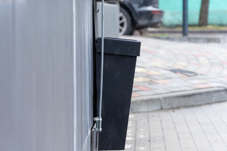 A black waste container, securely attached to a wall beside a sidewalk, enhances cleanliness in the...