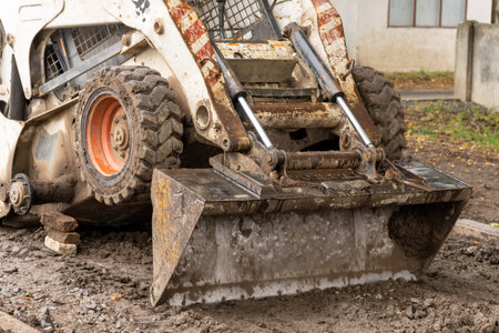 A skid steer loader with a dirt-filled bucket actively works on a muddy construction site, preparing...