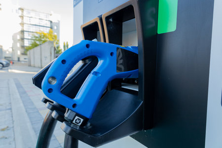 A blue fuel nozzle is positioned in a fuel pump at a contemporary gas station, surrounded by sleek buildings and sunlight.の写真素材