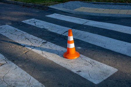 A bright orange traffic cone sits on a cracked crosswalk, marking a pedestrian area in a busy urban environment during dawn.の写真素材