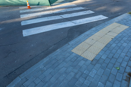 A view of a crosswalk featuring textured yellow paving, surrounded by gray bricks, and partially obstructed by construction barriers in daylight.の写真素材