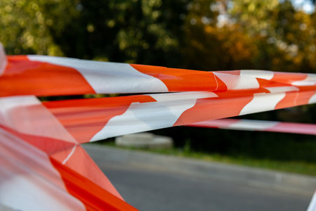 Bright orange and white barrier tape stretches across a path in a park, signaling a restricted area under vibrant trees during midday.の写真素材