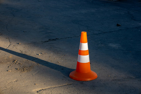 A brightly colored traffic cone stands on a sunlit road surface, indicating nearby construction or maintenance work is taking place.の写真素材