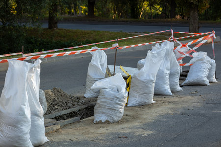 Sandbags and barriers surround an excavation area on a city road during late afternoon, indicating ongoing construction work.の写真素材