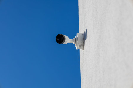 Modern security camera monitoring an outdoor area. White surveillance device fixed to a textured...
