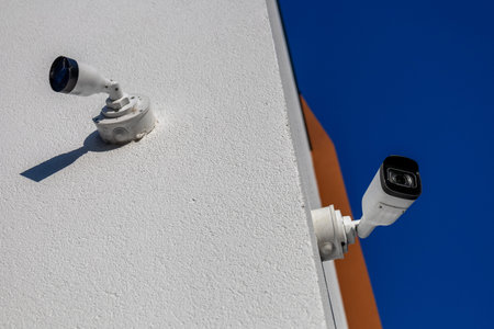 Two exterior security cameras are installed on a white wall, monitoring the area under a clear blue...