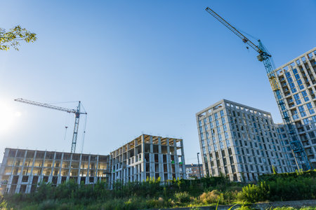 Cranes work above a busy construction site, where new residential buildings are being built alongside greenery under a clear blue sky.の写真素材