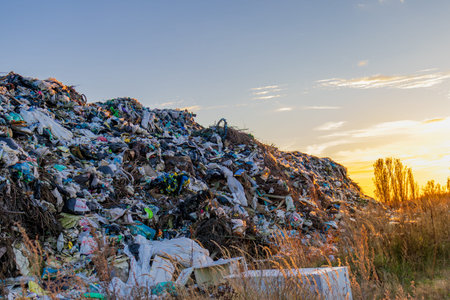 Garbage and plastic waste pile up in a landfill while the sun sets in the background, showcasing the...