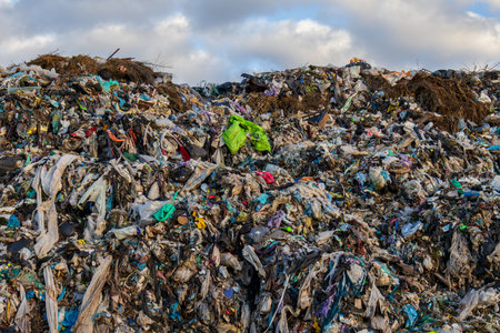 A large landfill site displays a chaotic mix of plastic waste, garbage, and organic matter under a...