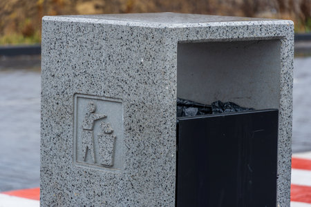 A sturdy trash bin made of stone is placed in a park, showing a symbol for proper waste disposal and filled with black trash bags.の写真素材