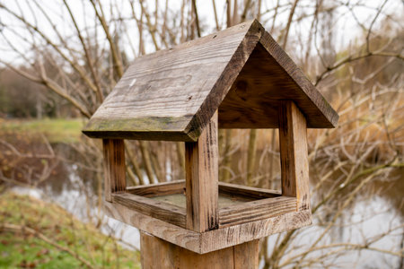 A rustic birdhouse sits on a wooden post near a tranquil pond, framed by leafless trees and an autumn landscape.の写真素材