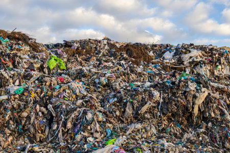 A large landfill site displays heaps of garbage including plastic and fabric waste under a cloudy...