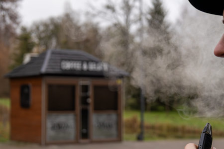 A person exhales vapor from a device while standing near a wooden kiosk in a park during autumn...